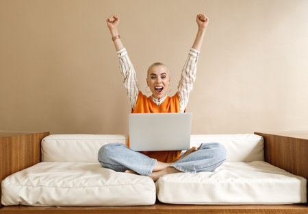 Happy businesswoman freelancer with trendy hairstyle and glasses raising arms and working at laptop screen with smile after finishing project while sitting on sofa in home officeの写真素材