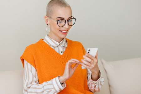 Young beautiful woman with very short hair sitting on sofa and using smartphone, texting message or surfing internet while relaxing at home. female with cell phoneの写真素材