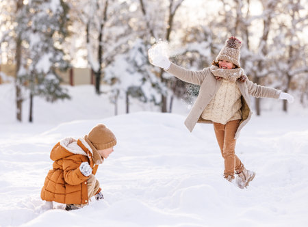 Happy children in warm clothes throwing snowballs at each other and laughing with joy, spending time on fresh air in wintertime, two overjoyed kids playing with snow in winter park on cold frosty dayの写真素材