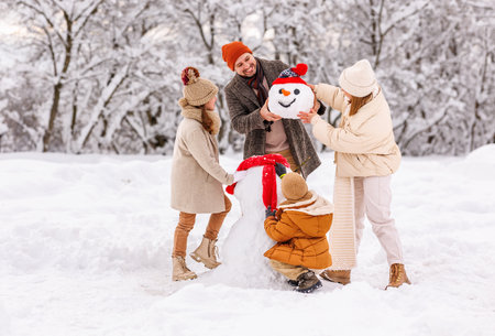 Happy parents and children gathering in snow-covered park together sculpting funny snowman from freshly fallen snow. Father, mother and two kids playing outdoors in winter forest. family active holidaysの写真素材