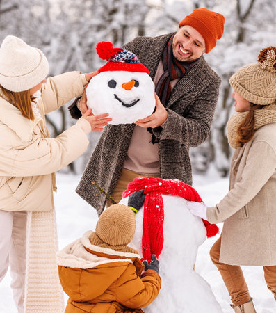 Happy parents and children gathering in snow-covered park together sculpting funny snowman from freshly fallen snow. Father, mother and two kids playing outdoors in winter forest. family active holidaysの写真素材