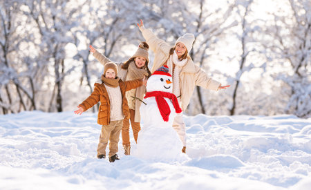 Young mom and two kids aughing merrily and raising hands up while posing around cheerful funny snowman in red hat and scarf, kids playing with mother in snowy park outdoorsの写真素材