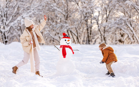 Happy overjoyed family mother and little son play snowballs outdoors in wintertime while standing near snowman, mom and kid laughing, having fun and playing with snow in snowy winter parkの写真素材