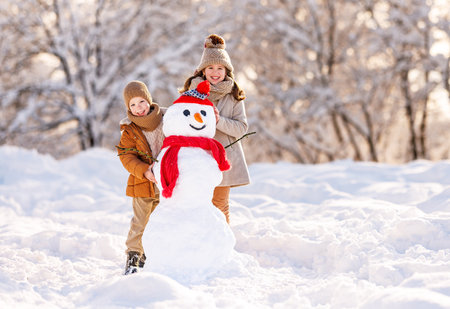 Happy children little brother and sister dressed in warm stylish clothes standing in winter snowy park broadly smiling while proudly posing near snowman made by them, kids playing with snow outdoorsの写真素材