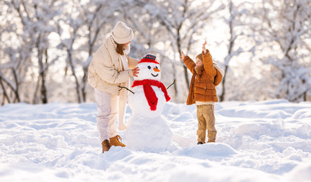 Cute little boy making snowman with mom in winter park. Happy young family mother and son in warm clothes building snow figure while playing and actively spending time on fresh frosty air in natureの写真素材
