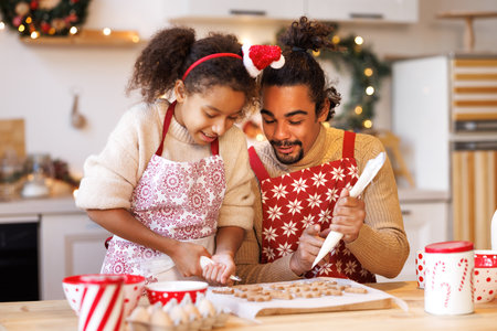 Happy african american family father and daughter in aprons making Christmas cookies together while cooking in kitchen at home, smiling girl helping dad to decorate xmas gingerbreads during winter holidaysの写真素材