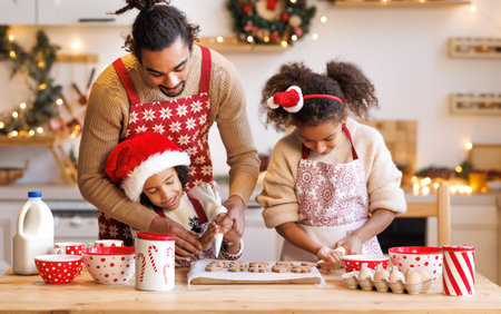 Happy african american family father and two kids in festive outfit making Christmas cookies together in kitchen at home during winter holidays, dad decorating xmas gingerbreads with small children siblingsの写真素材
