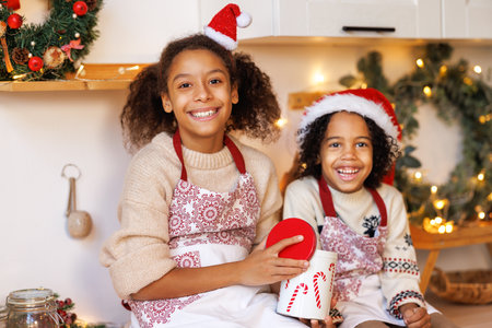 Happy funny african american kids on Christmas eve, girl and boy laugh and eat cookies that they baked together in cozy kitchen at homeの写真素材