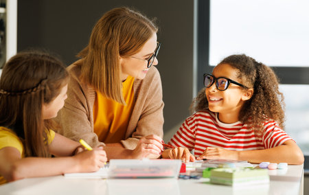 Smart woman teacher in glasses helping students to schoolwork during lesson in light classroom at schoolの写真素材