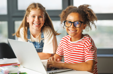 Positive kids pupils school girls happy smiling, sitting at desk and looking at camera during online lesson on laptopの写真素材