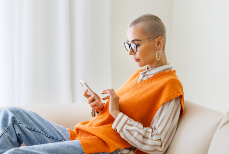 Young beautiful woman with very short hair sitting on sofa and using smartphone, texting message or surfing internet while relaxing at home. female with cell phoneの写真素材