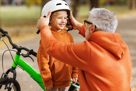 Happy familygrandfather puts on grandson helmet for safe cycling in autumn park in natureの写真素材