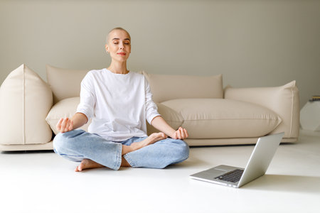 Calm young woman with very short hair sitting in Lotus pose on floor and meditating with lclosed eyes with laptop in morning at homeの写真素材