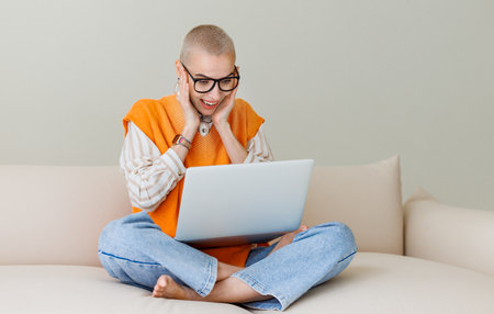 Beautiful woman freelancer with short hair works, makes video call and happily gestures while sitting on sofa and working on laptop during work from homeの写真素材