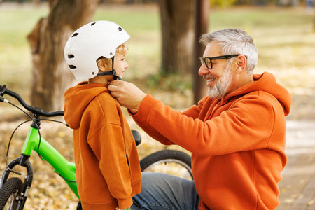 Happy familygrandfather puts on grandson helmet for safe cycling in autumn park in natureの写真素材