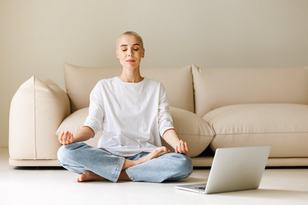 Calm young woman with very short hair sitting in Lotus pose on floor and meditating with lclosed eyes with laptop in morning at homeの写真素材