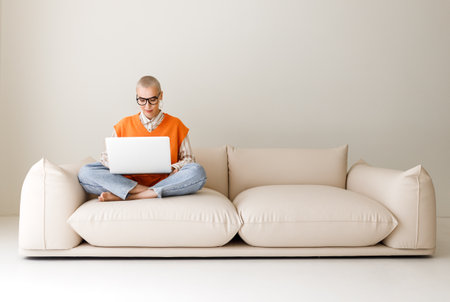 Beautiful woman freelancer in glasses with short hair works, while sitting on sofa and working on laptop during work from home in daytimeの写真素材