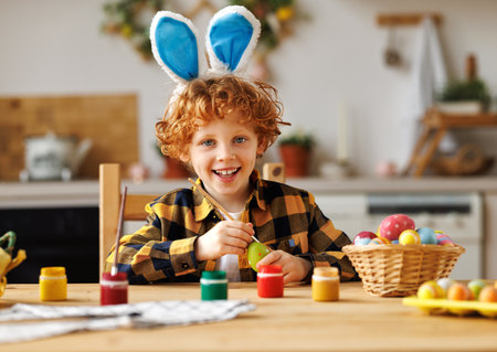 Cheerful cute child boy sitting at table and painting eggs while preparing for Easter celebration and looking at cameraの写真素材