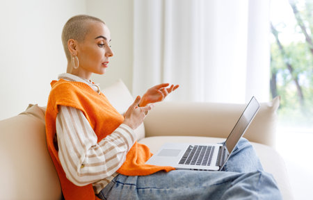 Beautiful woman freelancer with short hair works, makes video call and focused gestures while sitting on sofa and working on laptop during work from homeの写真素材