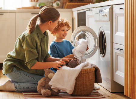 Cheerful family: kid son helping mother at linen in basket while doing laundry near washing machine in flight kitchen in weekend at homeの写真素材