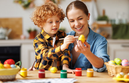 Easter Family Traditions. Loving young mother teaching happy little kid soon to dye and decorate eggs with paints for Easter holidays while sitting together at kitchen tableの写真素材