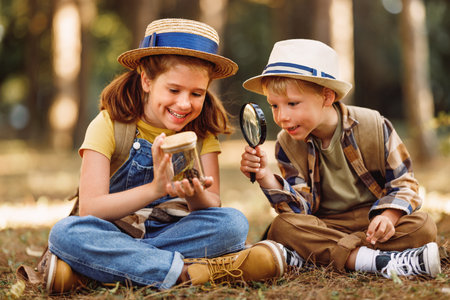 Two little children boy and girl with backpacks looking examining plants and insects in a jar through magnifying glass while exploring forest nature and environment on sunny day during outdoor ecology school lessonの写真素材
