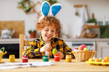Cheerful cute child boy sitting at table and painting eggs while preparing for Easter celebrationの写真素材