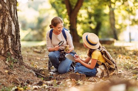 Child girl and smiling female teacher with backpacks looking examining plants and insects in a jar through magnifying glass while exploring forest nature and environment on sunny day during outdoor ecology school lessonの写真素材