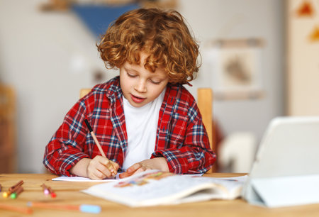 distance education. Smiling ginger child schoolboy studying online on tablet at home, sitting at table and communicating with teacher through video call on computerの写真素材