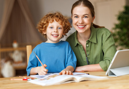 Smiling teacher or mother and little schoolboy doing exercises in copybook while studying remotely via tablet, while doing assignment together and studying remotely at homeの写真素材