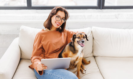 Happy young woman spending time with her pet beloved dog at home sitting on couch and browsing news in tabletの写真素材