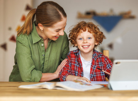 Smiling teacher or mother and little schoolboy doing exercises in copybook while studying remotely via tablet, while doing assignment together and studying remotely at homeの写真素材