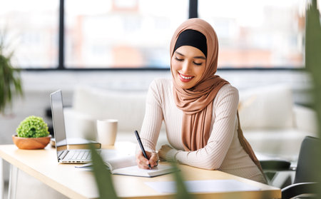 beautiful young smiling muslim woman in traditional religious hijab works remotely on laptop from homeの写真素材