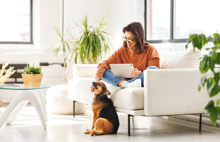 Happy young woman spending time with her pet beloved dog at home sitting on couch and browsing news in tabletの写真素材