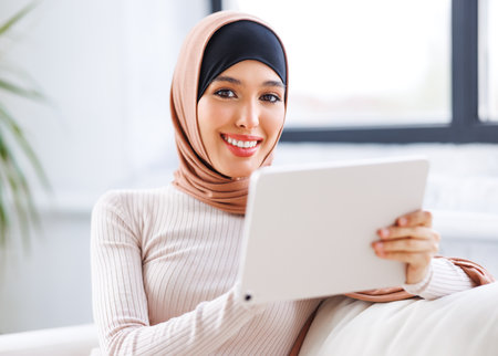 beautiful young smiling muslim woman in traditional religious hijab uses tablet while sitting on sofa at homeの写真素材