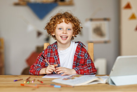 distance education. Smiling ginger child schoolboy studying online on tablet at home, sitting at table and looking at camera through video call on computerの写真素材