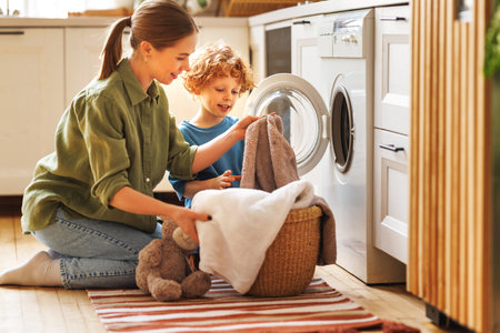 Cheerful family: kid son helping mother at linen in basket while doing laundry near washing machine in flight kitchen in weekend at homeの写真素材
