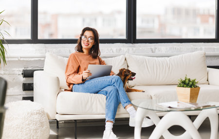 Happy young woman spending time with her pet beloved dog at home sitting on couch and browsing news in tabletの写真素材