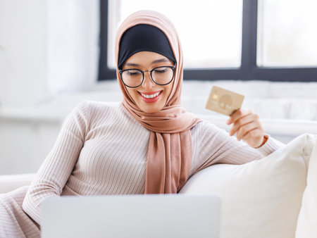 beautiful young smiling muslim woman in traditional religious hijab is doing online shopping while sitting on sofa at homeの写真素材