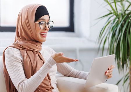 beautiful young smiling muslim woman in traditional religious hijab uses tablet smiling, gesticulating and talking to online while sitting on sofa at homeの写真素材