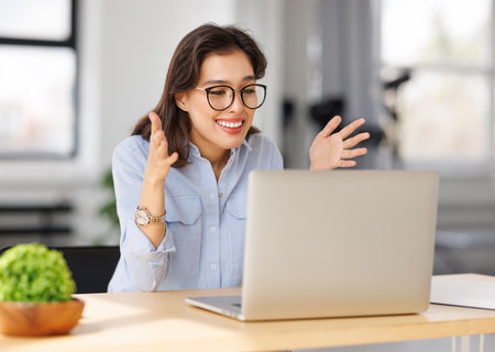 Joyful happy young female entrepreneur in glasses smiling and gesticulating while sitting at desk in home office and talking to online coworkers during online meetingの写真素材