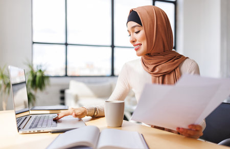 beautiful young smiling muslim woman in traditional religious hijab works remotely on laptop from homeの写真素材