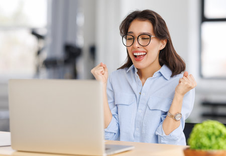 Joyful young business woman freelancer entrepreneur in glasses smiling and rejoices in victory while sitting at desk and working at laptop screen after finishing project in home officeの写真素材