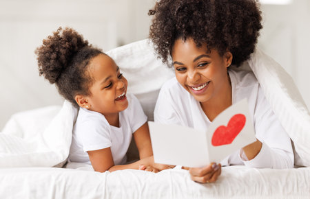 Cute little girl daughter congratulating his mom happy african american woman with Mother's day, giving her handmade greeting postcard with red heart at home. family holidays conceptの写真素材