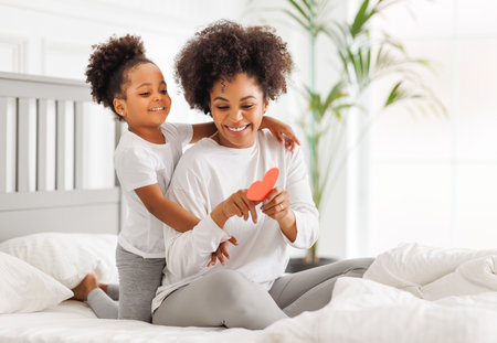 Cute little girl daughter congratulating his mom happy african american woman with Mother's day, giving her handmade greeting postcard with red heart at home. family holidays conceptの写真素材