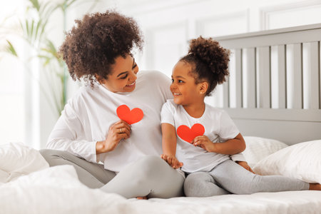Cute little girl daughter congratulating his mom happy african american woman with Mother's day, giving her handmade greeting postcard with red heart at home. family holidays conceptの写真素材