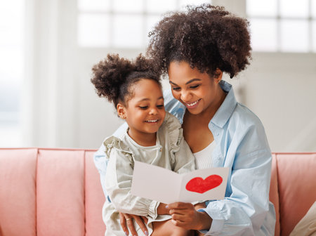 Cute little girl daughter congratulating his mom happy african american woman with Mother's day, giving her handmade greeting postcard with red heart at home. family holidays conceptの写真素材