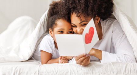 Cute little girl daughter congratulating his mom happy african american woman with Mother's day, giving her handmade greeting postcard with red heart at home. family holidays conceptの写真素材