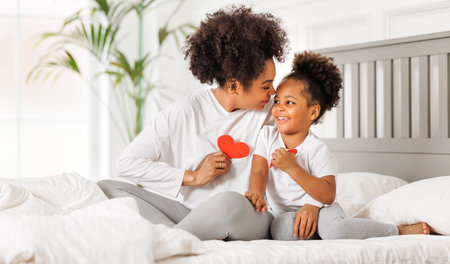 Cute little girl daughter congratulating his mom happy african american woman with Mother's day, giving her handmade greeting postcard with red heart at home. family holidays conceptの写真素材
