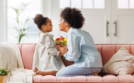Happy mother's day! Afro american family happy baby daughter congratulations mom on the holiday, hugs her and gives bouquet of flowers at homeの写真素材